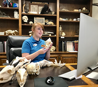 Person in a blue shirt holding a small object in front of a computer in a room with shelves displaying various items.