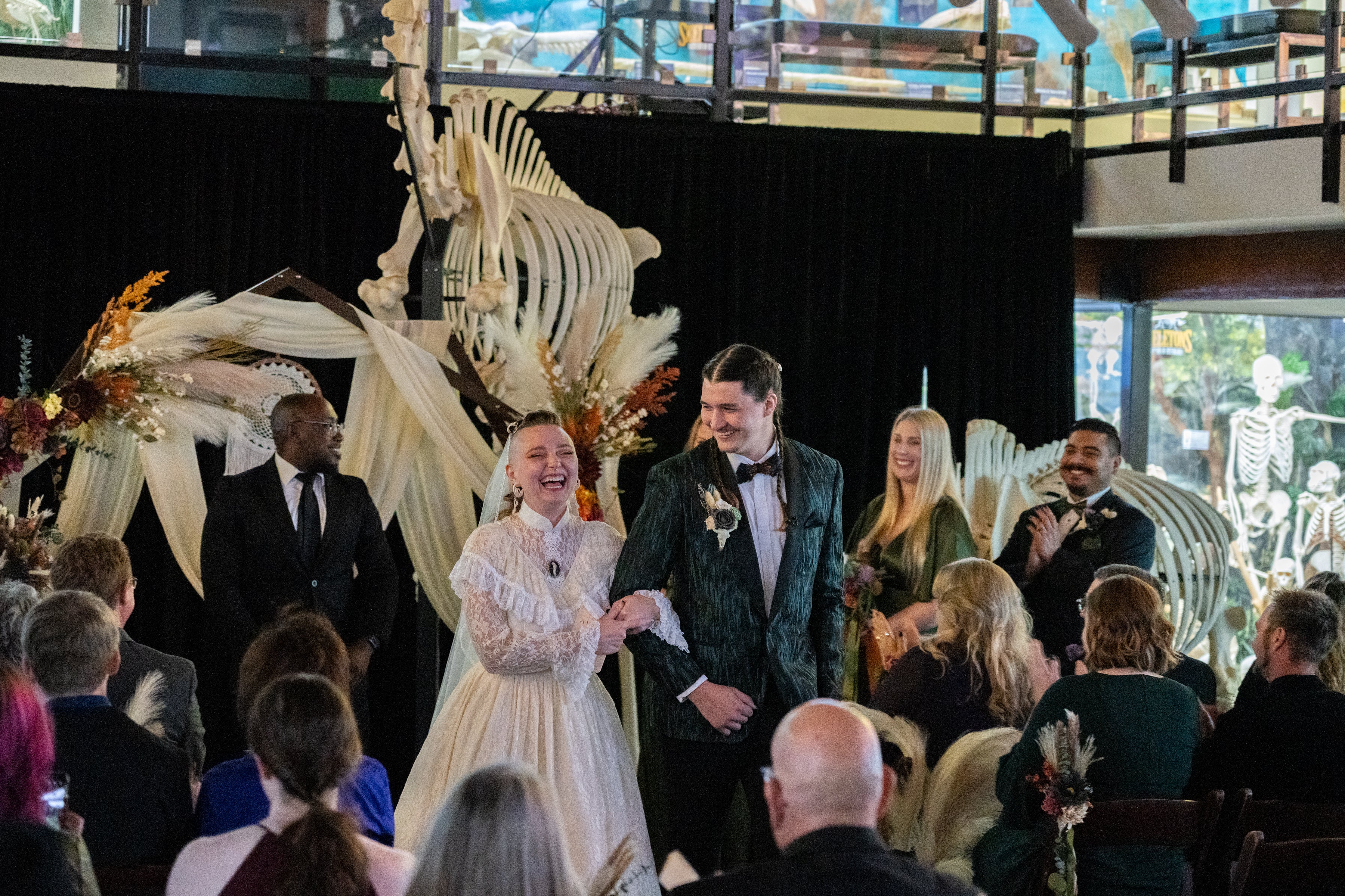 Wedding couple standing in front of a large skeleton with guests clapping.